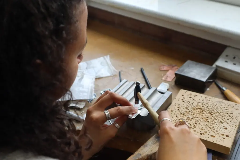 A young woman working at the small metals bench. She has silver rings on her fingers and is hammering another silver ring.